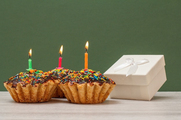 Tres deliciosos cupcakes de cumpleaños con glaseado de chocolate y caramelo, decorados con velas festivas encendidas y caja de regalo blanca sobre fondo verde. Feliz cumpleaños concepto mínimo.