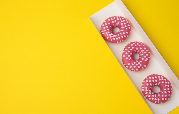 Três donuts redondos assados em esmalte rosa em uma caixa de papelão branca sobre um fundo amarelo, vista superior, espaço para cópia