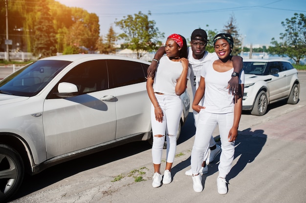 Tres elegantes amigos afroamericanos, usan ropa blanca contra dos autos de lujo. Moda callejera de jóvenes negros. Hombre negro con dos niñas africanas.