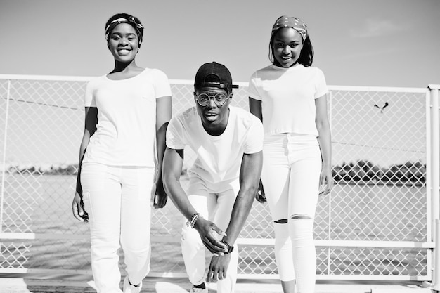 Tres elegantes amigos afroamericanos visten ropa blanca en el muelle en la playa contra la jaula Moda callejera de jóvenes negros Hombre negro con dos niñas africanas