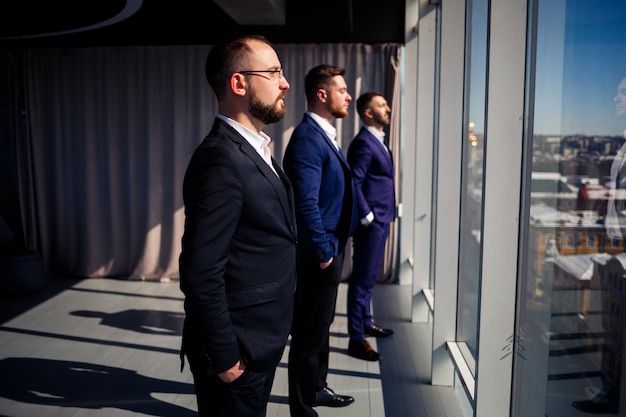 Foto três empresários de sucesso ficam em fila e olham pela janela com vista para a cidade em um grande arranha-céu