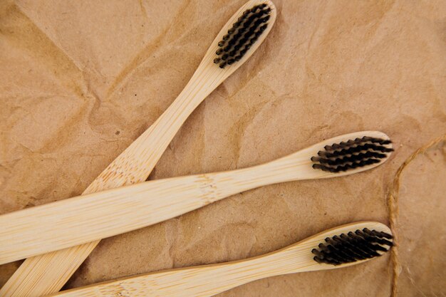 Três escovas de dente feitas de bambu natural estão sobre um pedaço de madeira, sobre um fundo de papel kraft marrom.