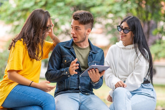 Tres estudiantes que estudian junto con una tableta digital sentados en un banco al aire libre