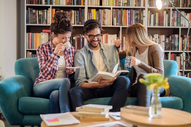 Tres estudiantes sonrientes que se sientan en el sofá en biblioteca y que miran el libro.