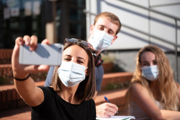 Tres estudiantes universitarios con máscaras faciales tomándose un selfie durante la pandemia del coronavirus o covid19