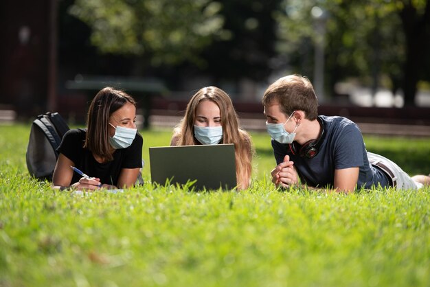 Tres estudiantes universitarios sonrientes con una computadora portátil tendida y charlando en un parque con una máscara facial