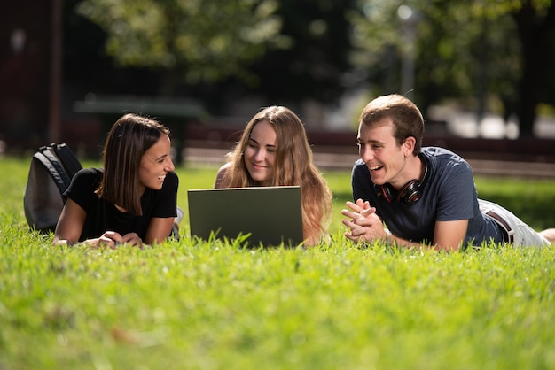 Tres estudiantes universitarios sonrientes usando una laptop tendida y charlando en un parque