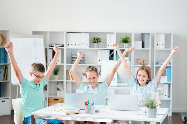 Tres extasiados compañeros de clase que mantienen sus manos levantadas mientras expresan alegría frente a sus computadoras portátiles