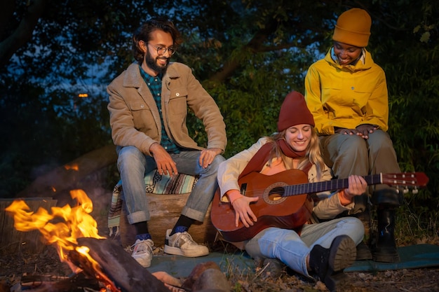 Tres felices y diversos amigos divirtiéndose tocando música y disfrutando de una fogata acampando en la naturaleza por la noche