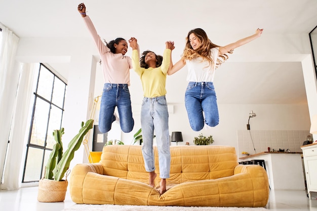 Três felizes amigas multiculturais pulando do sofá dentro de casa jovens mulheres alegres brincam e