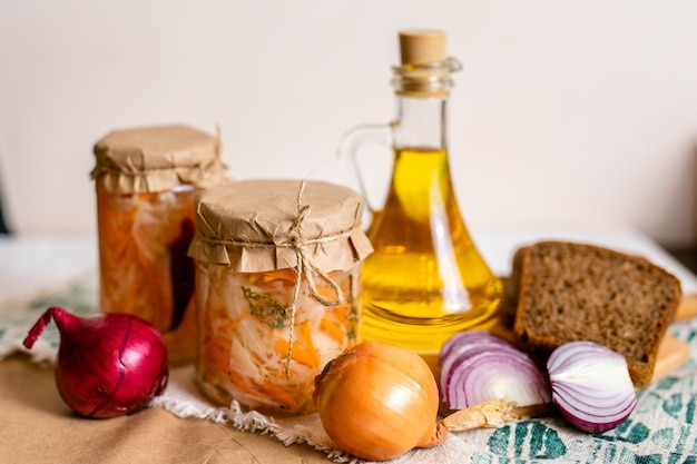 Tres frascos de chucrut y zanahorias en su propio jugo con especias y una botella de aceite, pan y cebolla, mesa de madera blanca. Plato tradicional fermentado casero de Rusia y Alemania