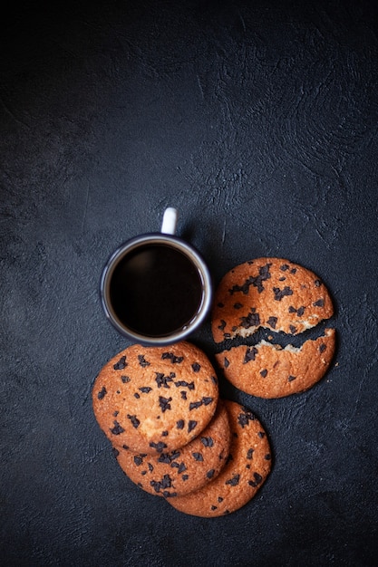 Tres galletas grandes y una taza de café sobre una superficie de hormigón negro. Una galleta se rompe en dos trozos Galletas con chocolate Imagen para inscripción