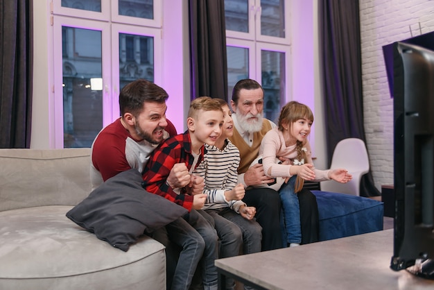 Tres generaciones atractivas de personas como padre, abuelo y nietos que se sientan en el cómodo sofá en casa y disfrutan de su tiempo libre viendo un partido de fútbol, gritando cuando el equipo anotó el balón.