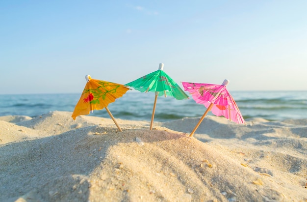 Três guarda-chuvas multicoloridos de coquetel de papel na praia de areia com céu azul