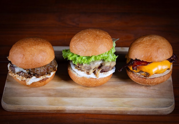 Foto três hambúrgueres colocados em uma tábua de madeira. hambúrgueres muito saborosos com queijo derretido, cheddar, bacon, alface, tomate, cebola e molho. burgers de carne artesanais feitos à mão.