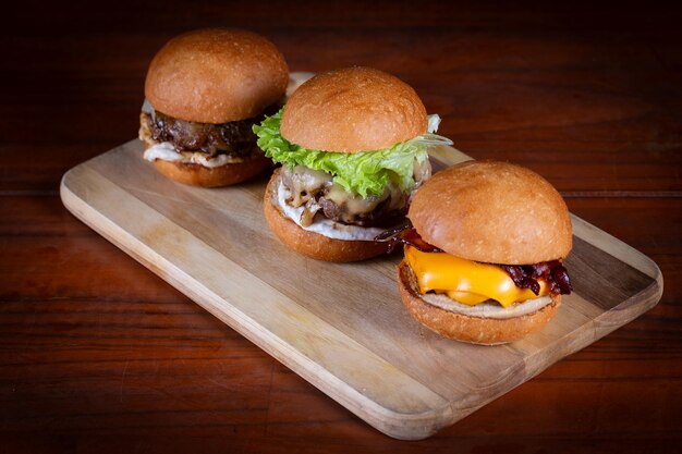 Foto três hambúrgueres colocados em uma tábua de madeira. hambúrgueres muito saborosos com queijo derretido, cheddar, bacon, alface, tomate, cebola e molho. burgers de carne artesanais feitos à mão.