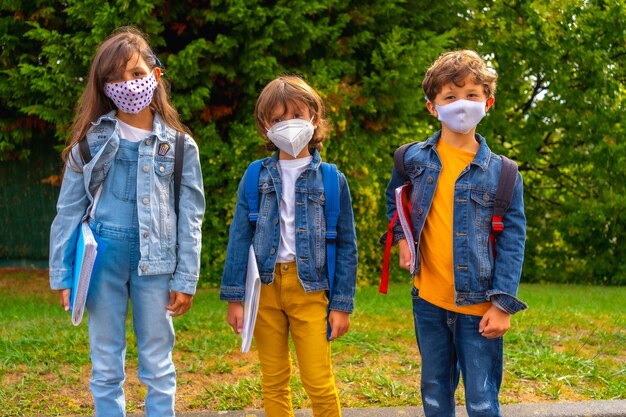 Tres hermanos con mascarillas listos para volver a la escuela. nueva normalidad, distancia social, pandemia de coronavirus, covid-19. esperando ir a la escuela con plantas verdes de fondo