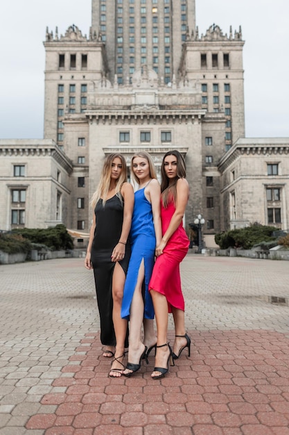 Tres hermosas y elegantes amigas esbeltas con un vestido de moda y zapatos de tacones se paran en la ciudad