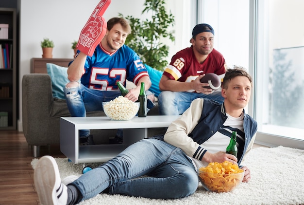 Tres hombres viendo partido con cervezas y bocadillos.