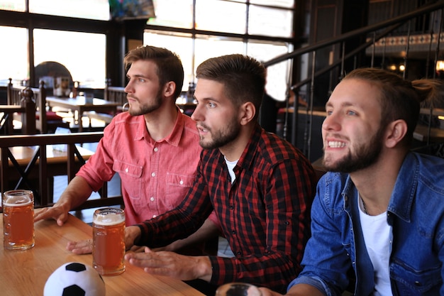 Três homens em roupas casuais estão torcendo pelo futebol e segurando garrafas de cerveja enquanto estão sentados no balcão de um bar.