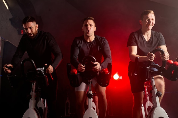 Três homens envolvidos em um simulador de bicicleta na academia, se exercitando em uma bicicleta ergométrica, isolados em um espaço esfumaçado com luz de néon escuro