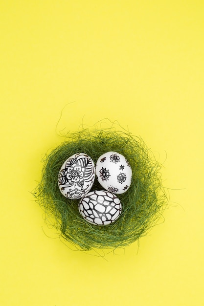 Tres huevos de Pascua están pintados en blanco y negro, abstracción, se encuentran en un nido verde. Huevos de Pascua pintados sobre un fondo amarillo. Endecha plana. Copia espacio