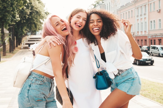 Três jovem linda sorridente hipster feminina em roupas da moda de verãoMulheres multirraciais despreocupadas sexy posando no fundo da ruaModelos positivos se divertindo em óculos de sol Alegre e feliz