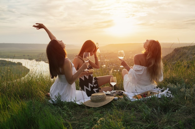 Tres jóvenes amigas felices con vestidos elegantes está haciendo un picnic en la colina al atardecer.