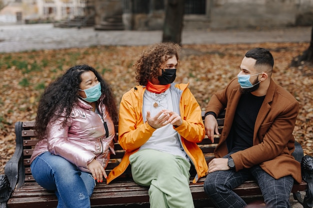 Tres jóvenes amigos multirraciales sentados en un banco de madera con máscaras médicas, sonriendo y hablando. Hombres y mujeres disfrutando de pasar tiempo juntos en el parque de otoño.