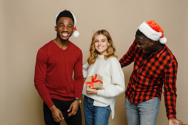 Tres jóvenes y hermosas amigas de diferentes nacionalidades con gorros de Papá Noel se alegran con una sorpresa y una caja de regalo.