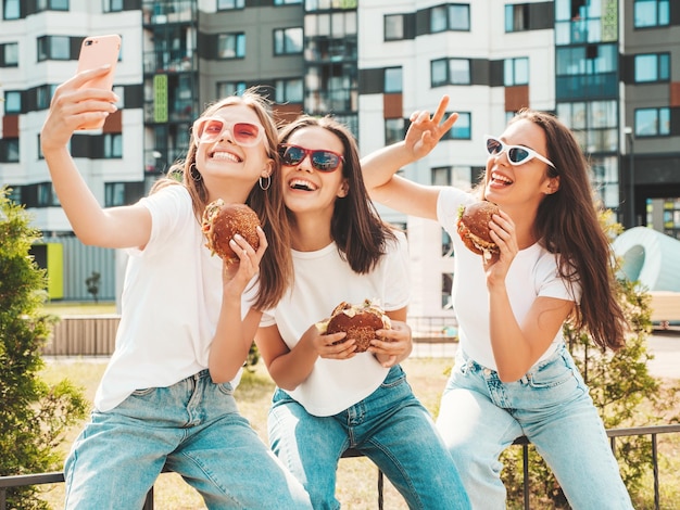 Tres jóvenes hermosas mujeres hipster sonrientes con ropa de verano de moda. Mujeres sexys y despreocupadas posando en la calle. Modelos positivos tomando selfie con gafas de sol. Sosteniendo una hamburguesa jugosa y comiendo una hamburguesa.