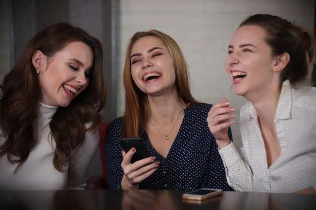 Tres jóvenes hermosas mujeres sentadas en un bar mirando el teléfono riendo