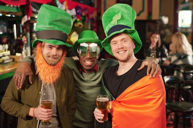 Tres jóvenes hombres interculturales alegres con sombreros verdes brindando con vasos de cerveza frente a la cámara contra los invitados sentados en la mesa en el pub