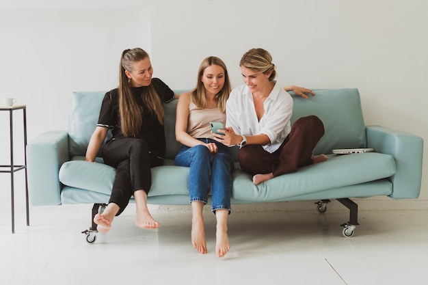 Tres jóvenes mujeres atractivas en casa en el sofá hablando, riendo y divirtiéndose, viendo fotos en el teléfono