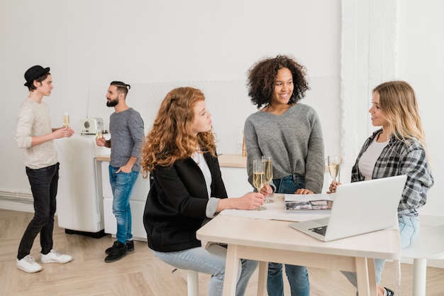 Tres jóvenes mujeres casuales con champán discutiendo algo por escritorio frente a la computadora portátil con dos chicos hablando