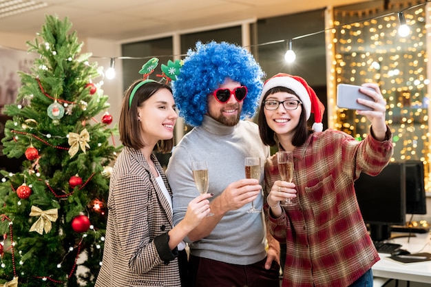 Tres jóvenes oficinistas con copas de champán mientras hacen selfie el día de Navidad