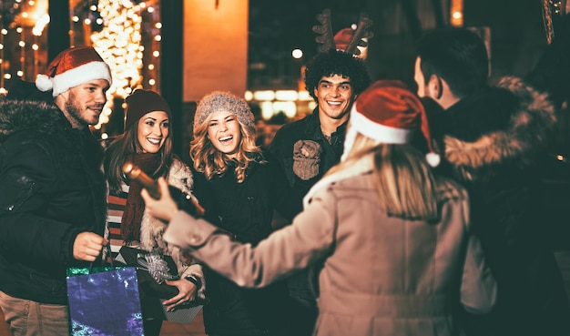 Tres jóvenes parejas alegres divirtiéndose en la fiesta de Navidad al aire libre en la calle de la ciudad por la noche y con muchas luces en el fondo.