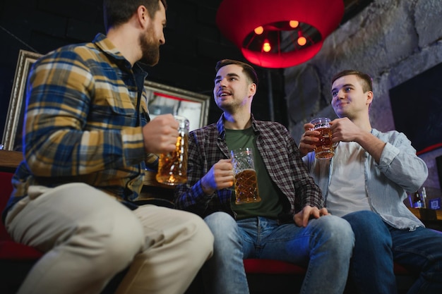 Tres jóvenes con ropa informal sonríen sosteniendo botellas de cerveza mientras están parados cerca del mostrador del bar en el pub