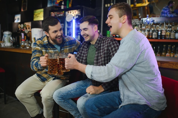 Tres jóvenes con ropa informal sonríen y tocan vasos de cerveza juntos mientras están sentados en el mostrador del bar en el pub