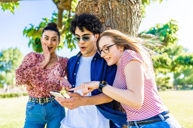 Três jovens amigos no parque da cidade com rostos surpresos, apontando o smartphone, aproveitando as redes sociais no celular. jovens se divertindo juntos na natureza com tecnologia de conexão de internet wi-fi