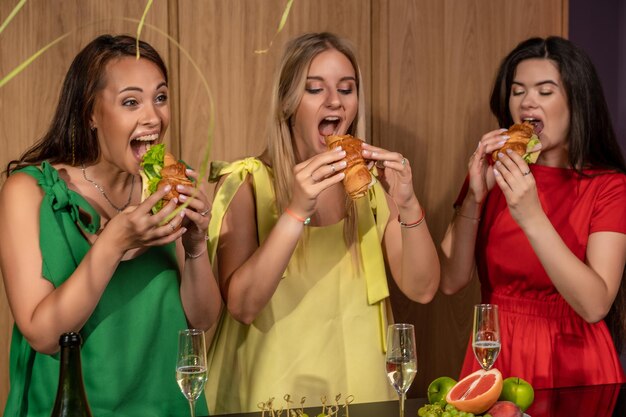 Três jovens despreocupadas se divertindo juntas na festa em casa com champanhe desfrutando de deliciosos sanduíches de croissant com verduras e presunto