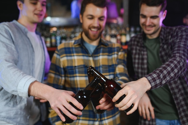 Três jovens em roupas casuais estão sorrindo e batendo copos de cerveja enquanto estão sentados no balcão do bar no pub