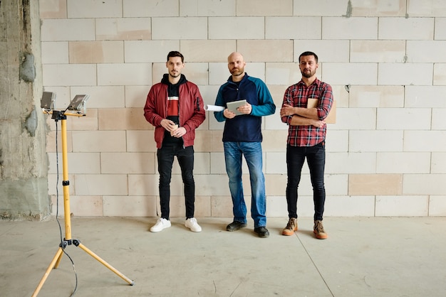 Três jovens engenheiros ou construtores em trajes de trabalho alinhados ao longo da parede de um prédio inacabado na frente da câmera durante o trabalho