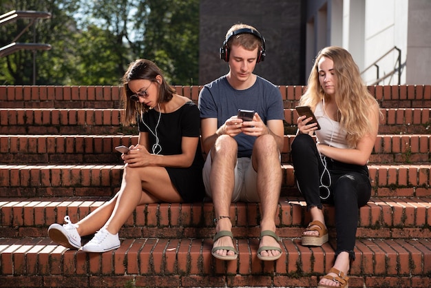 Três jovens estudantes sentados nas escadas e concentrados em seus smartphones