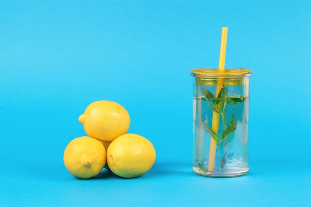 Tres limones y un vaso de agua con menta y limón sobre un fondo azul.