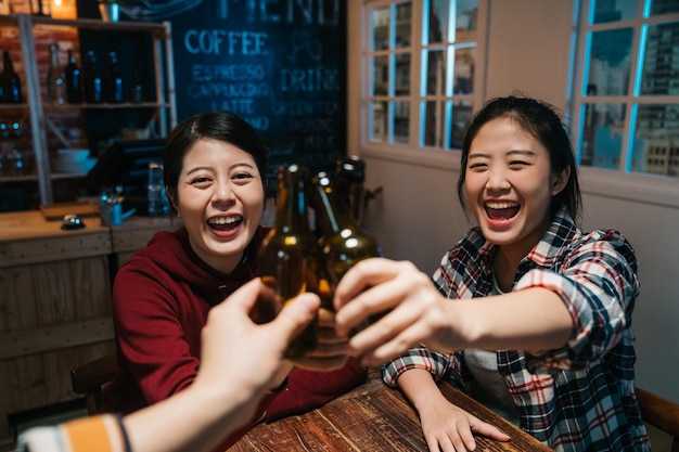 três lindas amigas tinindo garrafas de cerveja e sorrindo enquanto descansava no pub noturno. grupo de alegria de jovens felizes comemorando alegre rindo sentado no bar escuro da meia-noite.