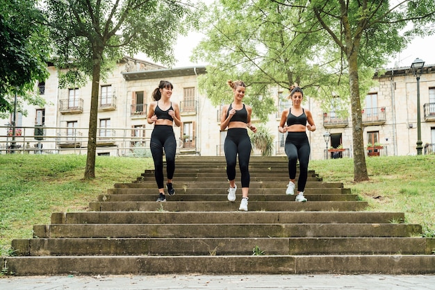 Três lindas mulheres descendo as escadas de um parque com muitas árvores da cidade, todas as três vestidas com roupas esportivas pretas