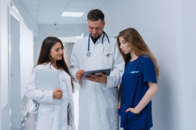 Tres médicos están parados en el pasillo de la clínica y discutiendo el diagnóstico del paciente. El médico jefe muestra a sus colegas el programa de tratamiento en la tableta.