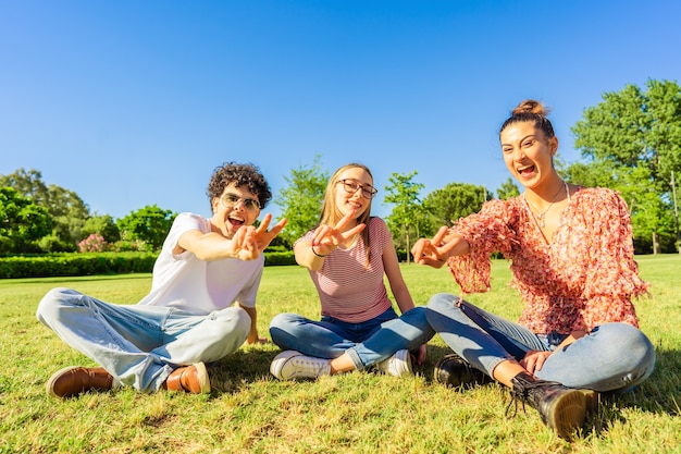 Três melhores amigos do jovem estudante sentado na grama no parque da cidade, mostrando o sinal de vitória com dois dedos, olhando para a câmera. Conceito de unidade e solidariedade na juventude. Geração feliz sorrindo na natureza