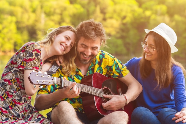 Três melhores amigos tocando violão juntos durante a viagem de férias Grupo de amigos felizes se divertindo à beira do rio Estilo de vida pessoas amizade e música